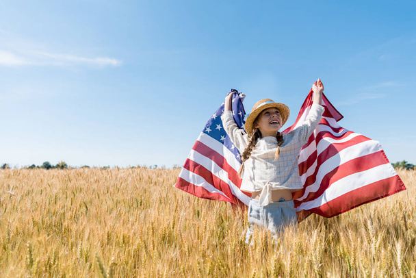 χαρούμενο παιδί σε ψάθινο καπέλο κρατώντας αμερικάνικη σημαία στο χρυσό χωράφι  - Φωτογραφία, εικόνα