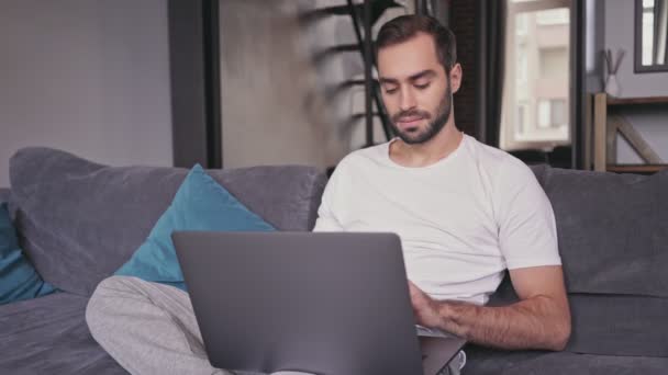 Hombre barbudo guapo serio usando computadora portátil mientras está sentado en el sofá en casa
 - Metraje, vídeo
