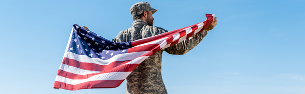 mavi gökyüzüne karşı Amerikan bayrağı tutan şapka lı ve üniformalı askerin panoramik fotoğrafı  - Fotoğraf, Görsel