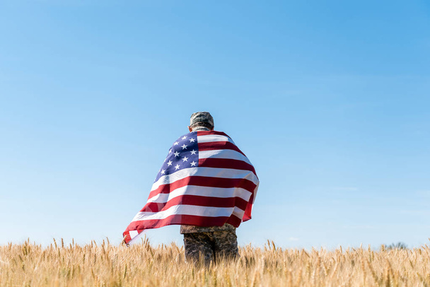 πίσω άποψη στρατιώτης σε Cap και ομοιόμορφη κρατώντας αμερικάνικη σημαία στο πεδίο  - Φωτογραφία, εικόνα