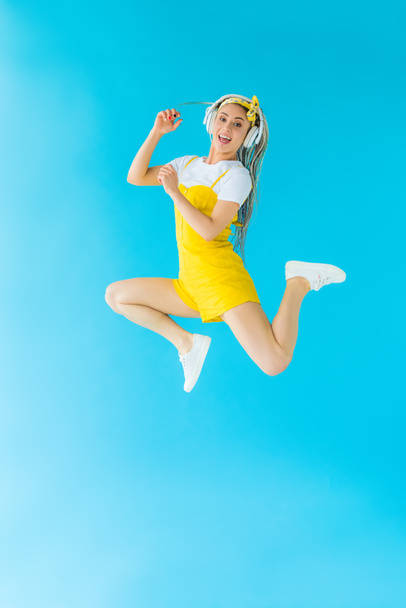 opgewonden meisje met dreadlocks in hoofdtelefoon springen op Turquoise - Foto, afbeelding