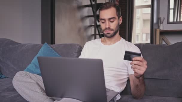 Carefree handsome bearded man pays the bills on laptop computer while sitting on sofa at home - Imágenes, Vídeo