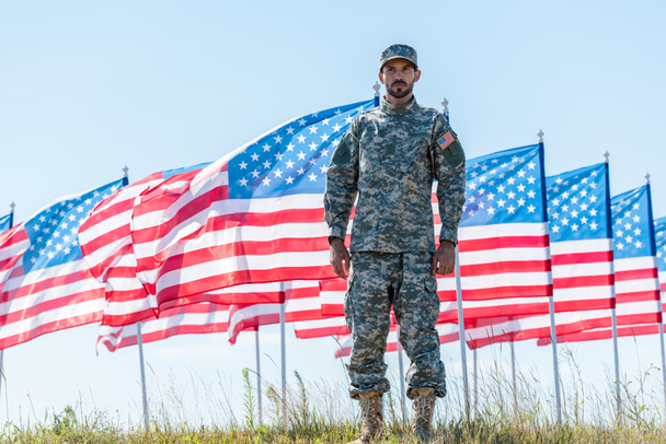 soldado bonito de uniforme em pé perto de bandeiras americanas e céu azul
  - Foto, Imagem