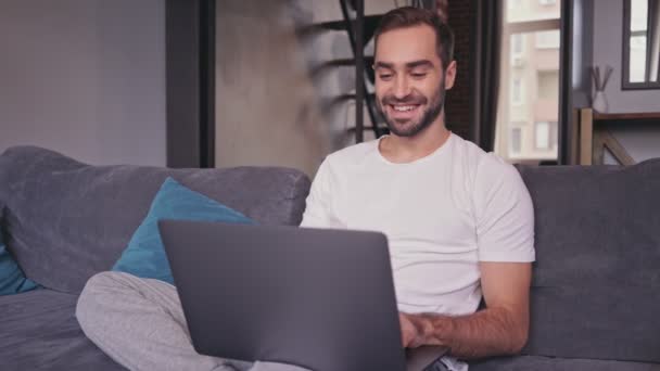 Carefree handsome bearded man using laptop computer while sitting on sofa at home - Metraje, vídeo