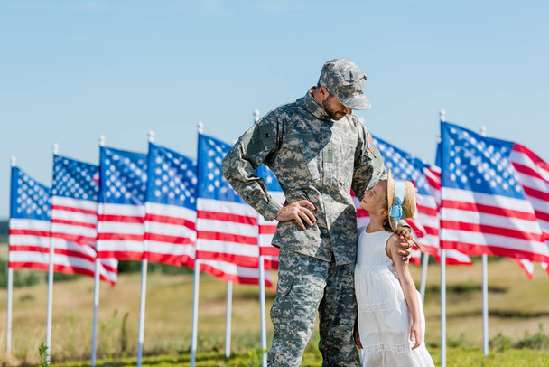 military man in cap looking at kid in straw hat near american flags with stars and stripes  - Fotografie, Obrázek