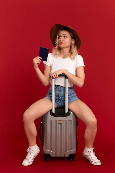 Surprised young woman with blod curly hair in a sundown hat sits on a grey luggage bag holding passport infront of a red background - Фото, изображение