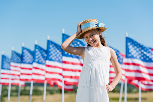 gelukkig kind staande in witte jurk en aanraken van stro hoed in de buurt van Amerikaanse vlaggen  - Foto, afbeelding