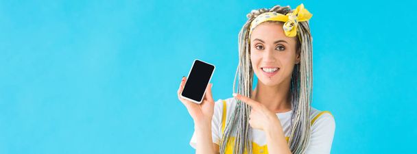 panoramic shot of girl with dreadlocks pointing with finger at smartphone with blank screen isolated on turquoise - Foto, imagen