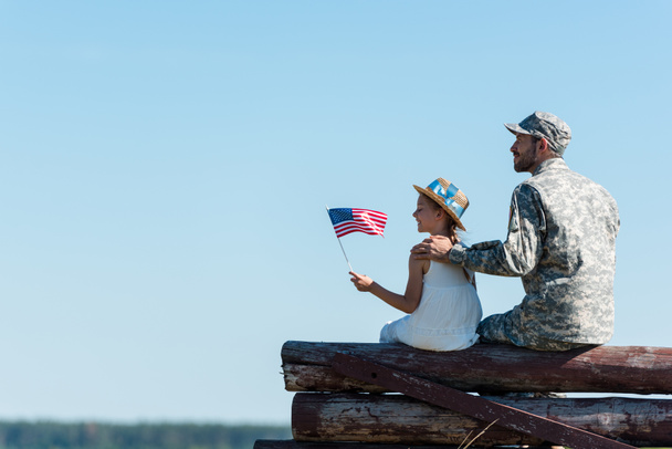 lindo niño sosteniendo bandera americana cerca de padre veterano mientras está sentado en valla
  - Foto, Imagen