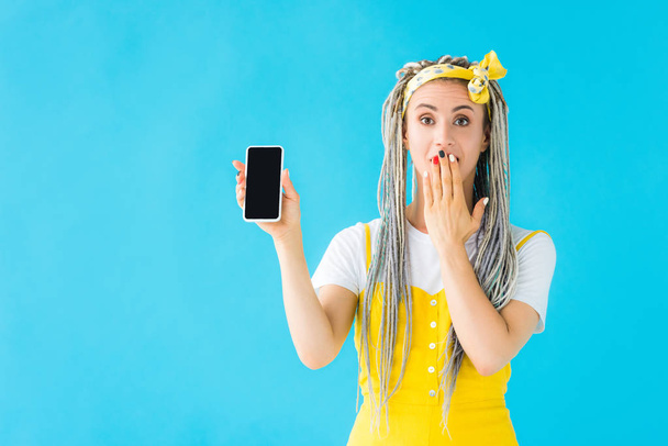 chica con rastas cubriendo la boca y mostrando smartphone con pantalla en blanco aislado en turquesa
 - Foto, Imagen
