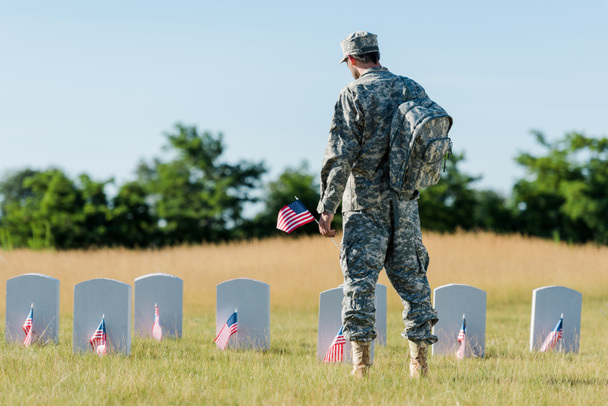 Rückansicht eines Soldaten mit amerikanischer Flagge in der Nähe von Grabsteinen auf dem Friedhof  - Foto, Bild