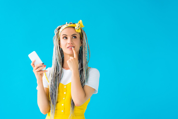 pensive girl with dreadlocks and finger on mouth holding smartphone isolated on turquoise - Fotoğraf, Görsel