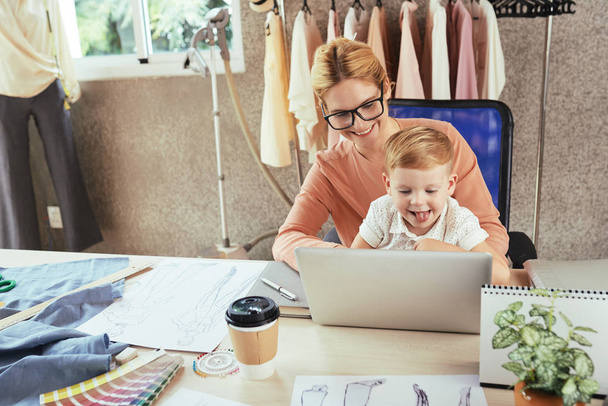 fröhliche Modedesignerin arbeitet am Laptop mit Sohn auf dem Schoß - Foto, Bild