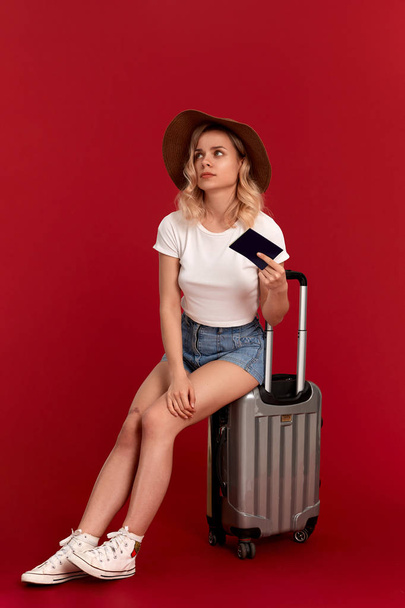 Bir günbatımı şapka blod kıvırcık saçlı yorgun genç kadın pasaport tutan gri bir bagaj çantası üzerinde oturur - Fotoğraf, Görsel