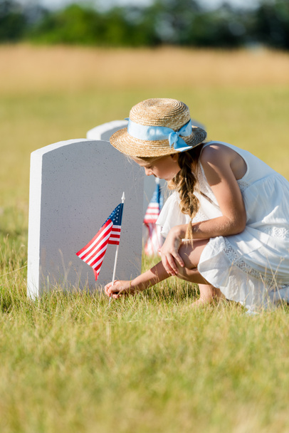 selective focus of adorable kid sitting near headstone with american flag in graveyard  - Photo, Image