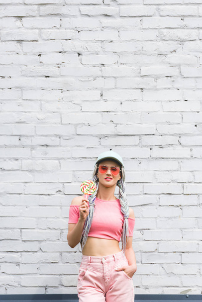 girl in hat and sunglasses holding lollipop near brick wall - Fotoğraf, Görsel