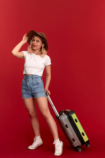 Happy young blond curly woman in a sundown hat holds grey luggage bag while standing infront of a red background - Fotó, kép