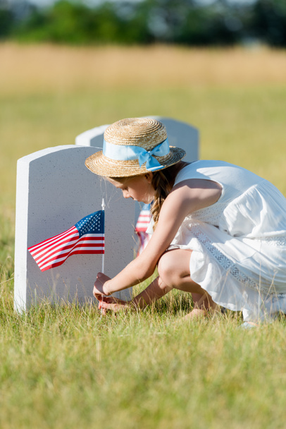 selective focus of adorable child sitting near headstone with american flag in graveyard  - Photo, Image