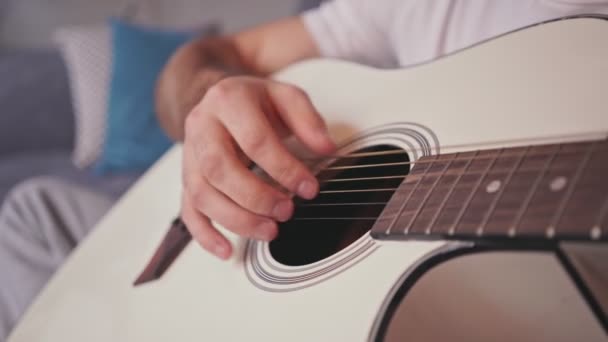 Close up θέα του νεαρού άνδρα που παίζει στην κιθάρα, ενώ κάθεται στον καναπέ στο σπίτι - Πλάνα, βίντεο