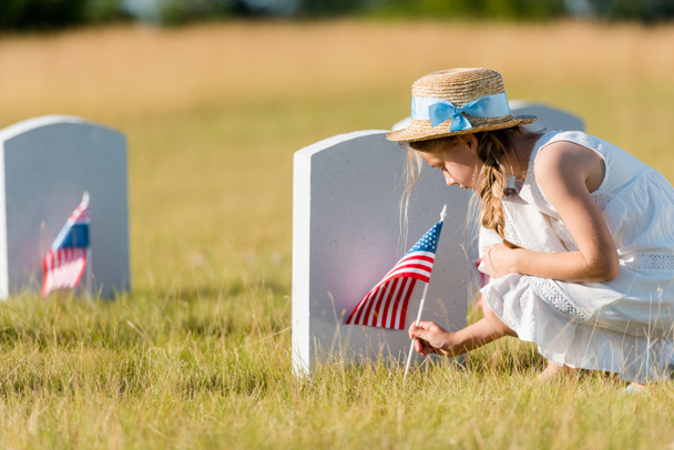 selective focus of adorable kid in straw hat sitting near headstone with american flag in graveyard  - Fotografie, Obrázek