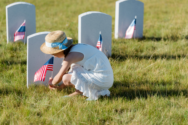 selective focus of  kid in straw hat sitting near headstone with american flag in graveyard  - Foto, Imagen