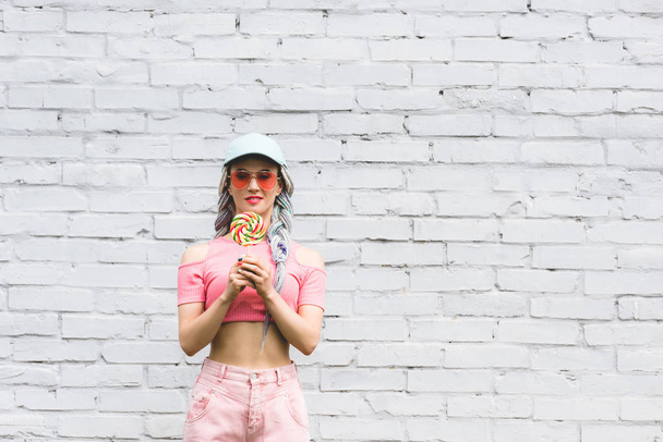 girl in hat and sunglasses holding lollipop near brick wall with copy space - Foto, immagini