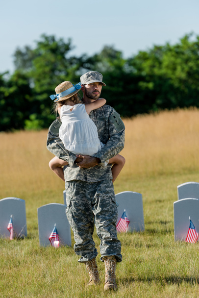 Militärmann mit Mütze hält ein Kind mit Strohhut in der Nähe von Grabsteinen mit amerikanischen Flaggen  - Foto, Bild
