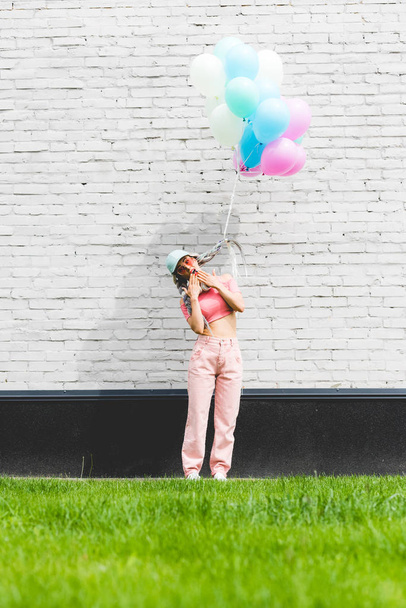 stylish girl with decorative balloons covering mouth near brick wall - Fotografie, Obrázek