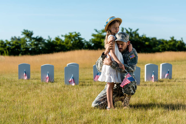 militar en uniforme abrazando niño cerca de lápidas en cementerio
  - Foto, Imagen