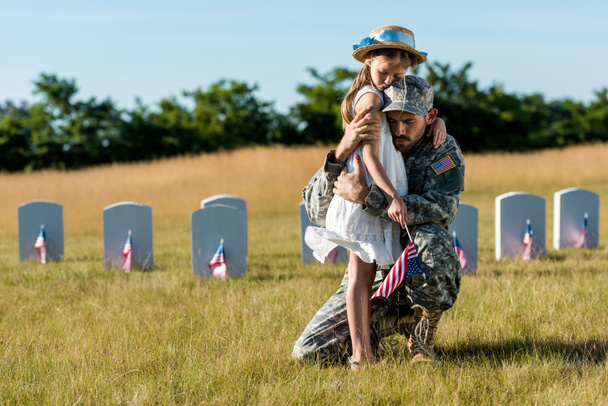 militar en uniforme abrazando hija cerca de lápidas en cementerio
  - Foto, Imagen
