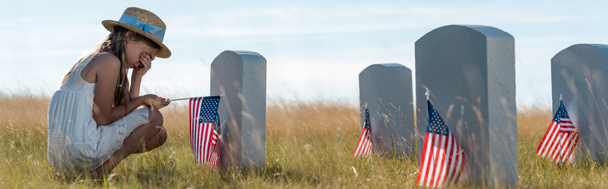 plano panorámico de niño en sombrero de paja cubriendo la cara mientras está sentado cerca de lápidas con banderas americanas
  - Foto, Imagen