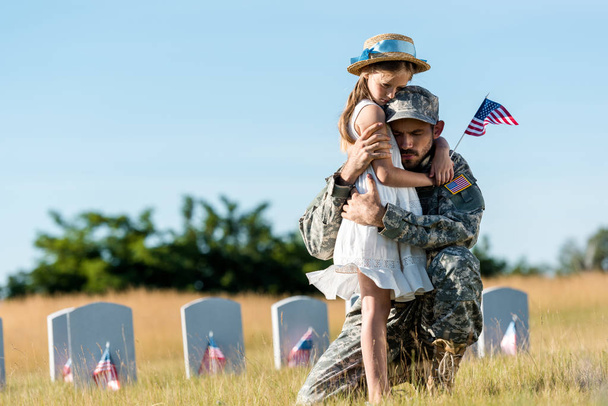 militar con los ojos cerrados abrazando hija cerca de lápidas en el cementerio
  - Foto, Imagen