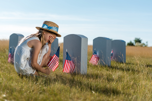 enfant en chapeau de paille couvrant le visage tout en étant assis près des pierres tombales avec des drapeaux américains
  - Photo, image