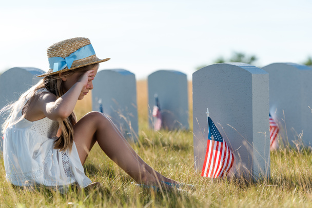 kid in straw hat touching face while sitting near headstones with american flags  - Photo, Image