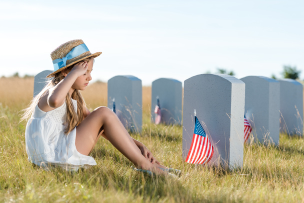 niño en sombrero de paja dando saludo mientras está sentado cerca de lápidas con banderas americanas
  - Foto, Imagen