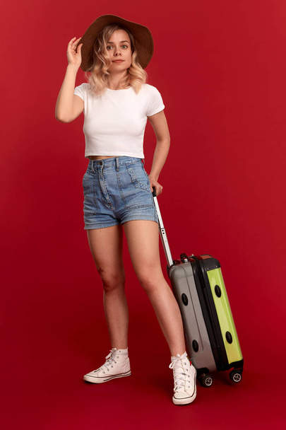 Щаслива молода блондинка кучерява жінка в сонячному капелюсі тримає сірий багаж, стоячи на передньому плані червоного фону
 - Фото, зображення