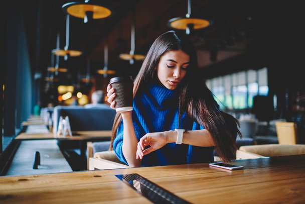 Caucasian beautiful hipster girl looking at wearable smartwatch while enjoying free time with coffee sitting at cafeteria at wooden table, attractive young woman checking notifications on wristwatch - Φωτογραφία, εικόνα