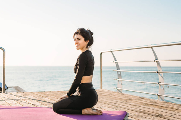 Smiling brunette young woman in a black jumpsuit rests after plank workout on a mat on pier at sunrise.  - Photo, Image