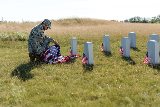 enfoque selectivo de soldado en uniforme y gorra con bandera americana mientras está sentado en el cementerio
  - Foto, Imagen