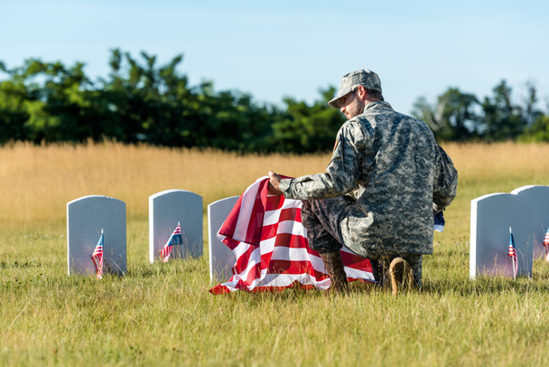 hombre en uniforme militar y gorra sosteniendo bandera americana mientras está sentado en el cementerio
  - Foto, Imagen