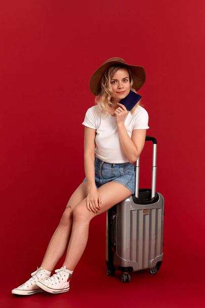 Happy young woman with blod curly hair in a sundown hat sits on a grey luggage bag holding passport infront of a red background - Photo, image
