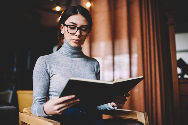 Young good looking hipster girl in trendy apparel holding notebook and reading interesting stories, concentrated attractive woman in eyewear enjoying favourite literature at university campus - Zdjęcie, obraz