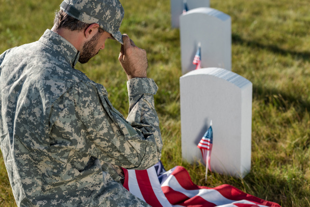 enfoque selectivo de soldado en camuflaje uniforme tocando la tapa mientras está sentado en el cementerio
  - Foto, Imagen