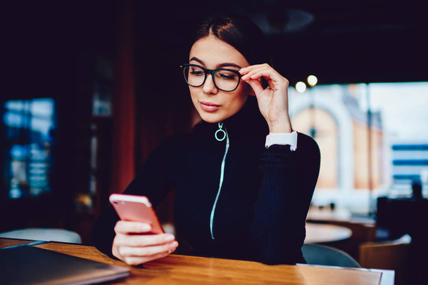 Attractive teenage hipster girl surfing internet using smartphone device connected to wifi, good looking brunette woman in spectacles sending message from cellphone and checking mail indoors - Photo, Image