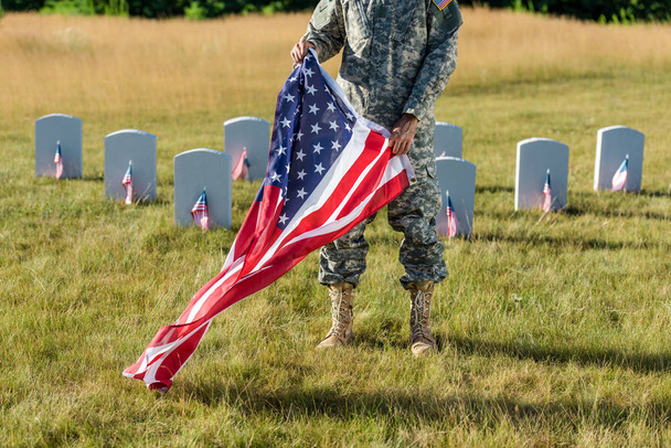 κομμένη θέα του ανθρώπου σε καμουφλάζ στολή κρατώντας αμερικάνικη σημαία και στέκεται στο νεκροταφείο  - Φωτογραφία, εικόνα