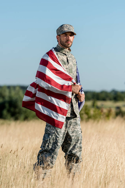 金色のフィールドでアメリカの旗を保持する迷彩制服と帽子をかぶった軍人  - 写真・画像