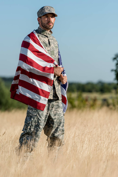 man in camouflage uniform and cap holding american flag in golden field  - Photo, Image
