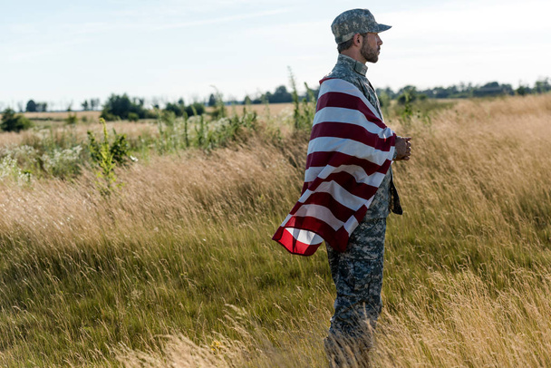 金色のフィールドでアメリカ国旗を掲げる迷彩ユニフォームを着た兵士の側面図  - 写真・画像