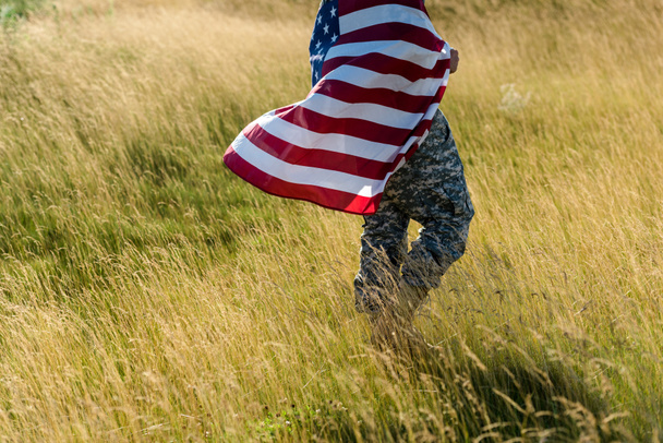 cropped view of soldier in camouflage uniform holding american flag in field  - Foto, afbeelding