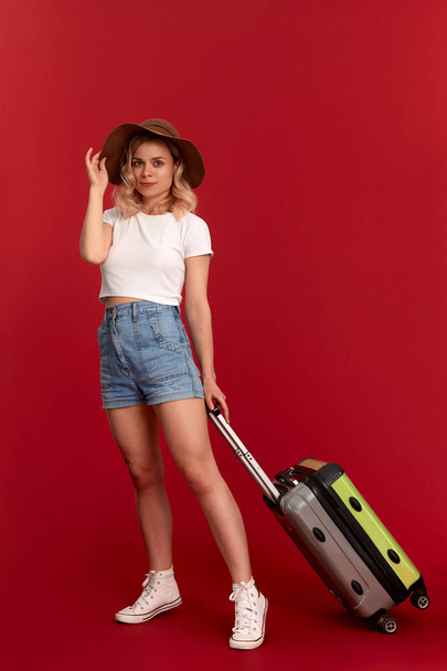 Happy young blond curly woman in a sundown hat holds grey luggage bag while standing infront of a red background - Fotoğraf, Görsel